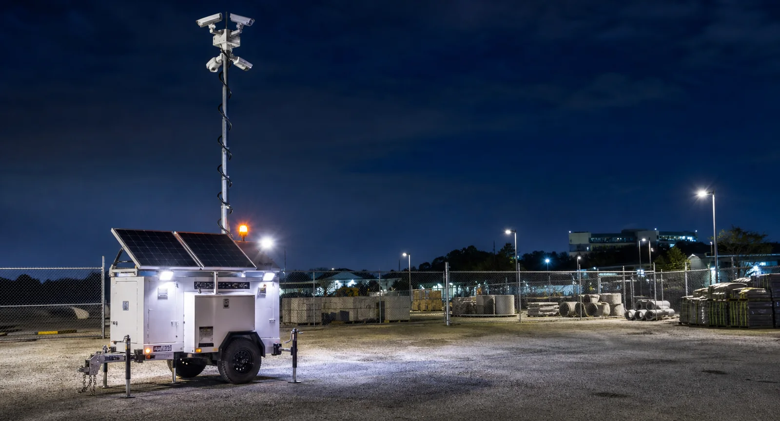 Illuminated mobile surveillance trailer monitoring a fenced material yard at night